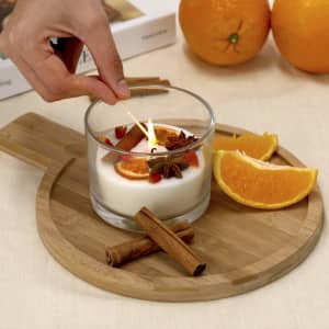 A glass candle with a lit wick sits on a wooden tray, surrounded by cinnamon sticks and orange slices.