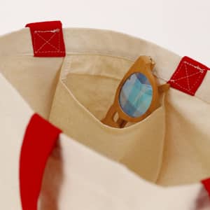 A tote bag with red handles, featuring a pocket that holds a pair of wooden sunglasses.