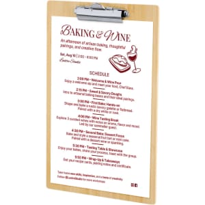 A wooden clipboard with a metallic clip, featuring a printed schedule for a baking and wine event.