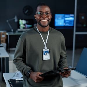 A person wearing a white lanyard with an ID badge stands in an office, holding a tablet and smiling.