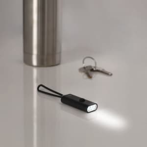 A black torch with a keyring attachment emits light, positioned near a drink bottle and a set of keys on a surface.