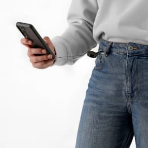 A person holds a smartphone with a cable attached to their jeans pocket. The phone case is dark and the jeans are blue.