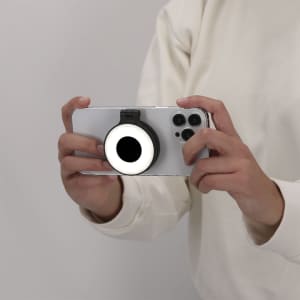 A person holding a smartphone with a black and white Cenob Ring Light attached to it. The background is neutral.