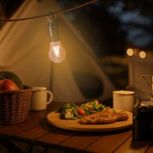 A Trapix Lamp hangs above a wooden table with a plate of food, surrounded by a basket, mugs, and a tent backdrop.