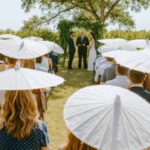 A group of people at a wedding ceremony, shaded by white, Asian-style umbrellas under a tree.