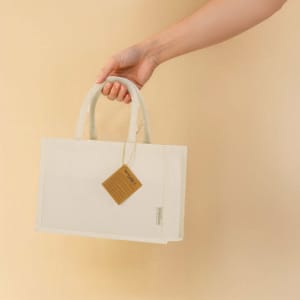 A hand holds a cream-coloured tote bag with short handles and a tag attached, featuring a logo.