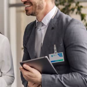 A man in a grey suit holds a tablet while wearing a badge holder on his lapel, with greenery in the background.