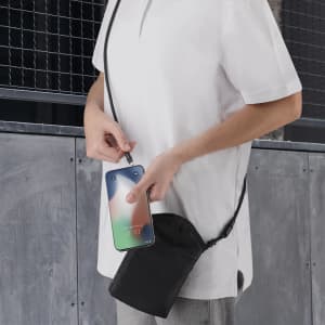 A person holds a black multipurpose bag with a phone visible inside. The bag has a strap and is worn crossbody.