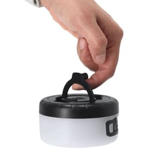 A hand holding a compact, round LED light with a black top and white base, featuring a carrying handle.