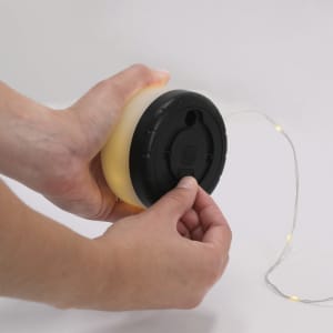 A hand holding a round, yellow LED light with a black base and a string of fairy lights attached.