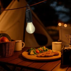 A warm white hanging lamp illuminates a campsite table with a plate of food, surrounded by a basket and drinks.