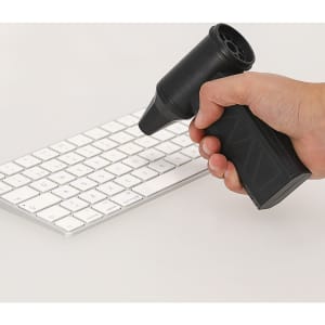 A black vacuum cleaner blower held in a hand, aimed at a white keyboard on a flat surface.
