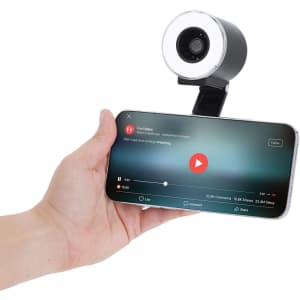 A hand holds a smartphone with a video playing, attached to a black and silver Tangix Fan Ring Light.
