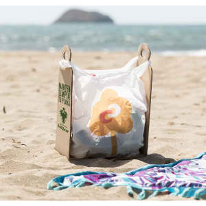 A cardboard bag holder in sand with a white trash bag featuring a flower design, by the beach near water.