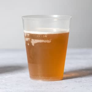A transparent plastic cup filled with amber-coloured liquid, showcasing a frothy top. The cup features a logo.