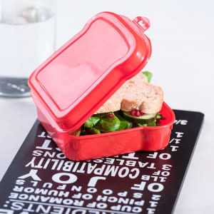 A red lunch box contains a sandwich with lettuce and cucumber, resting on a black textured surface.