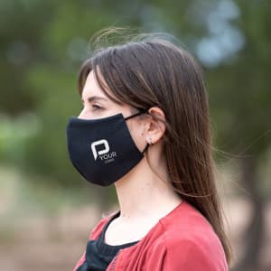 A black reusable hygienic mask displayed on a model, featuring a logo on the side, set against a natural background.