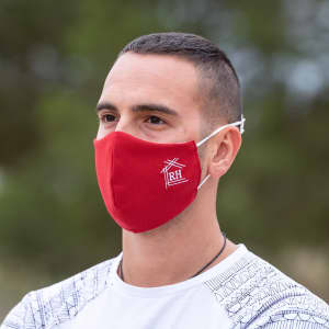 A red reusable hygienic mask worn by a person, featuring a logo on the side. The background is blurred greenery.