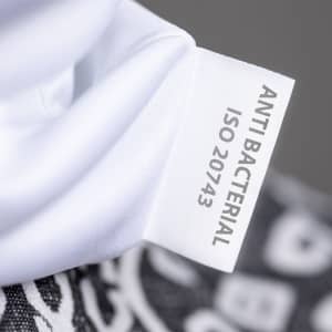 A white fabric label with "ANTI BACTERIAL ISO 20743" displayed, featuring part of a patterned neck warmer.