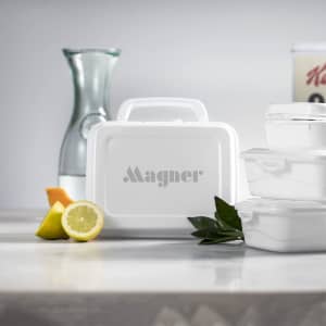 A white lunch box with a handle and logo, surrounded by citrus fruits and glass containers on a marble surface.