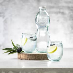 A drink bottle and two glasses of water with lemon and berries, set on a wooden board. The glassware is clear and stylish.