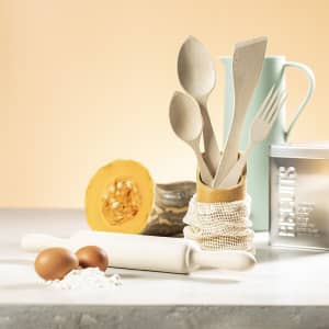 A wooden rolling pin rests beside eggs and flour, with kitchen utensils in a jar and a cut pumpkin in the background.