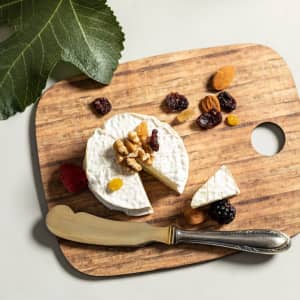 A wooden cutting board featuring a round cheese wheel, nuts, dried fruits, and a silver knife, with a leaf in the background.