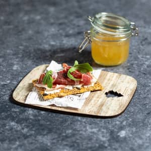 A wooden cutting board featuring a slice of crispbread topped with meat, herbs, and a small jar of preserves.