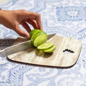 A wooden cutting board with a hand slicing lime, featuring a decorative blue tablecloth underneath.