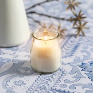 A white aromatic candle in a glass jar, placed on a decorative blue patterned surface with decorative elements nearby.