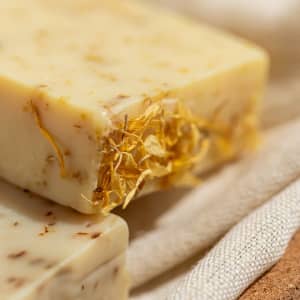 A close-up of a creamy bar of soap featuring dried flowers, resting on a soft, neutral fabric background.