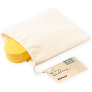 A yellow solid shampoo bar in a cream-coloured pouch, accompanied by a tag. The product is 100% vegan.