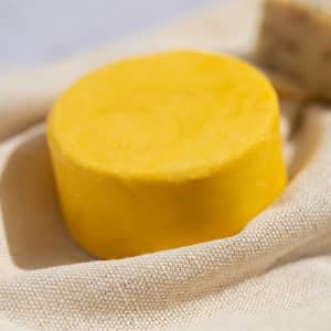 A round yellow solid shampoo bar sits on a beige cloth background. The product has a smooth surface and a simple design.