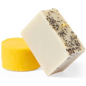 Two bars of soap: one yellow and round, one rectangular with a cream colour topped with black seeds. Both have a simple design.