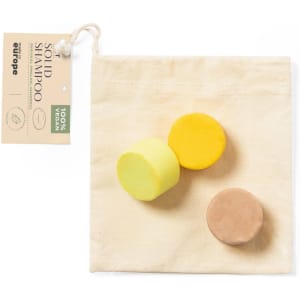 Three solid shampoo bars in yellow, light brown, and beige, packaged in a natural fabric bag with a branding label.