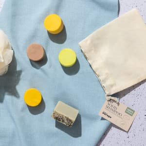 A collection of solid shampoo bars in yellow, green, brown, and a soap dish, accompanied by a light fabric pouch.