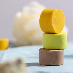 Three cylindrical shampoo bars stacked in yellow, green, and brown, set against a soft background.