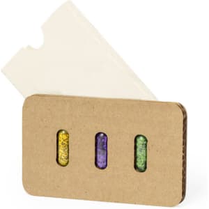 A set of three seed capsules in purple, green, and yellow, packaged in a brown cardboard holder with a white cover.