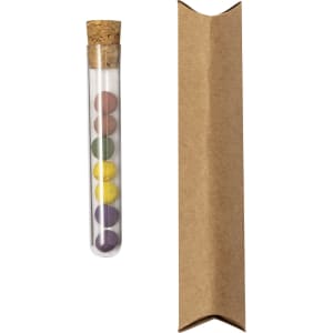 A clear test tube with a cork lid contains six colourful seed bonbons in shades of purple, yellow, and green, alongside a brown paper package.