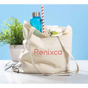 A cream tote bag with a logo, containing a blue drink bottle, a straw, and various items, alongside glasses and a plant.