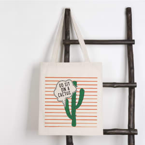 A cream tote bag hanging on a ladder, featuring a cactus illustration and orange striped background with a logo.