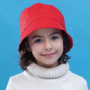 A red bucket hat on a young girl wearing a white turtleneck sweater, set against a blue background. The hat has a logo.