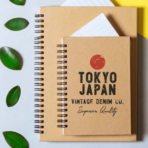 A set of two notebooks with brown covers, featuring spiral bindings and a logo on the front of the smaller notebook.