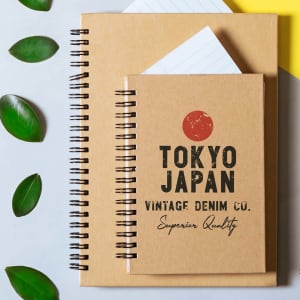 A duo of spiral-bound notebooks in kraft paper cover, featuring Japanese-inspired designs and a logo. Green leaves surround them.