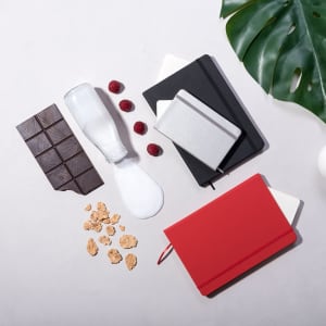 A variety of notepads in red, black, and grey, alongside chocolate, a milk bottle, raspberries, and granola.