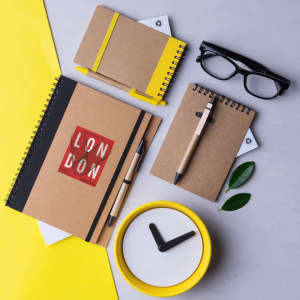 A set of notebooks in brown and yellow, a pen, glasses, and a yellow clock on a grey background.