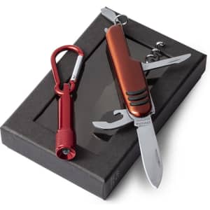 A red multi-tool with a knife and various attachments, accompanied by a red carabiner and a light, packaged in a black box.