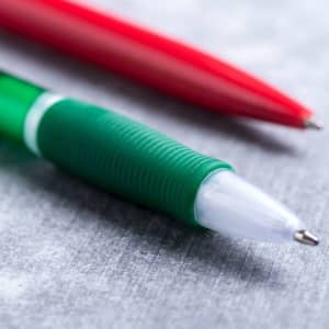 A green plastic pen with a grip and a red pen in the background, resting on a grey surface.