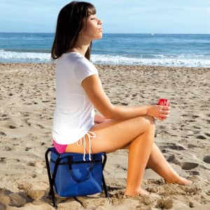 A blue chair cool bag sits on the sand, with a person wearing a white t-shirt and pink shorts holding a drink.