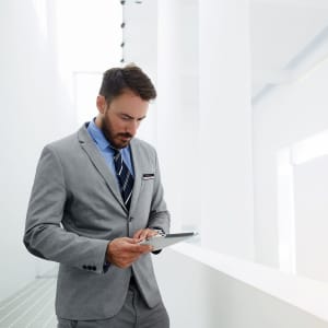 A man in a grey suit and striped tie stands looking at a tablet in a bright, modern space with white walls.